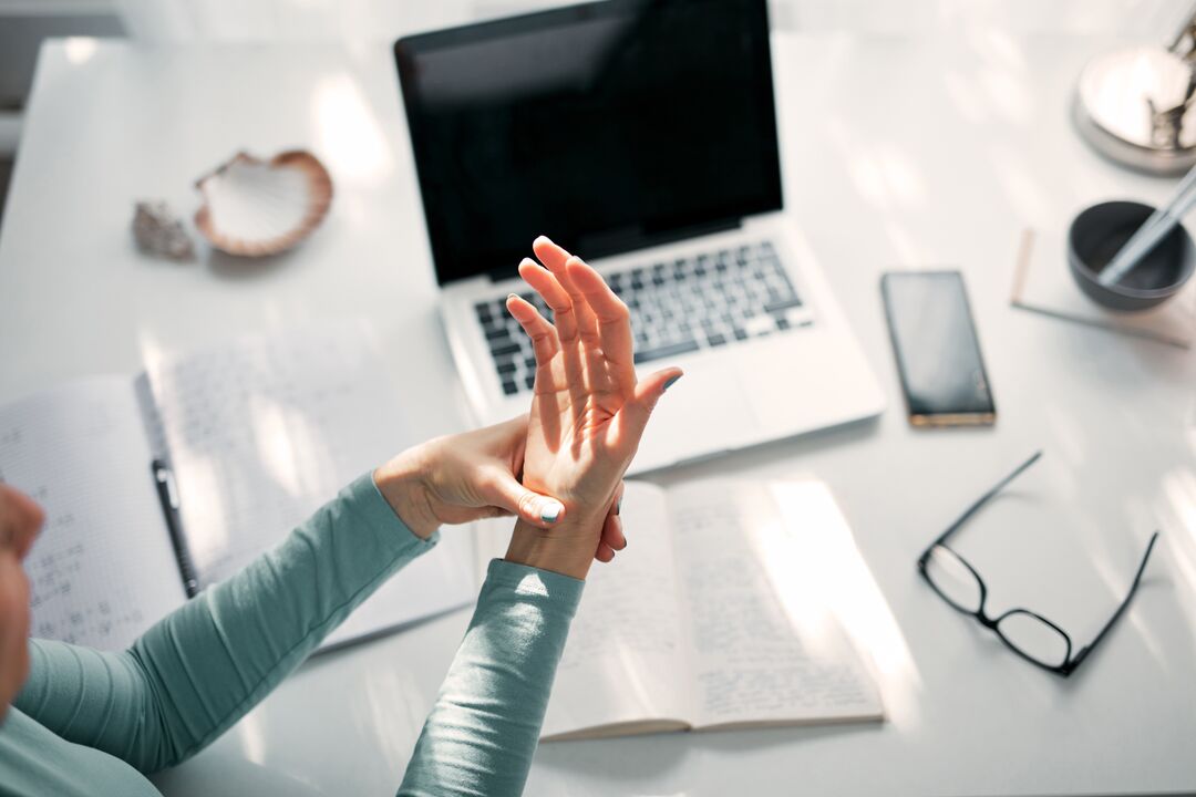 Person rubbing their wrist while sitting at a desk with a laptop and notebook.
