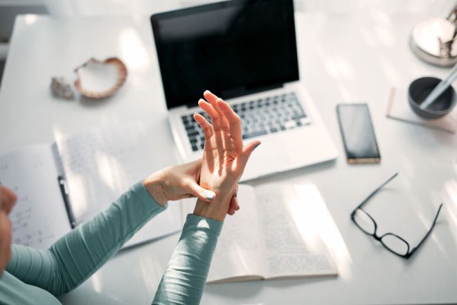 Person rubbing their wrist while sitting at a desk with a laptop and notebook.