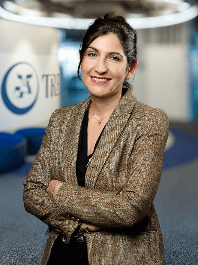 Leila Nobs, smiling, wearing a brown blazer, arms crossed in an office setting.