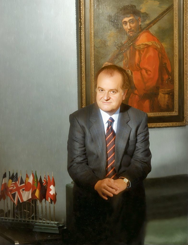 Man in a suit standing beside a painting of a man in red clothing.