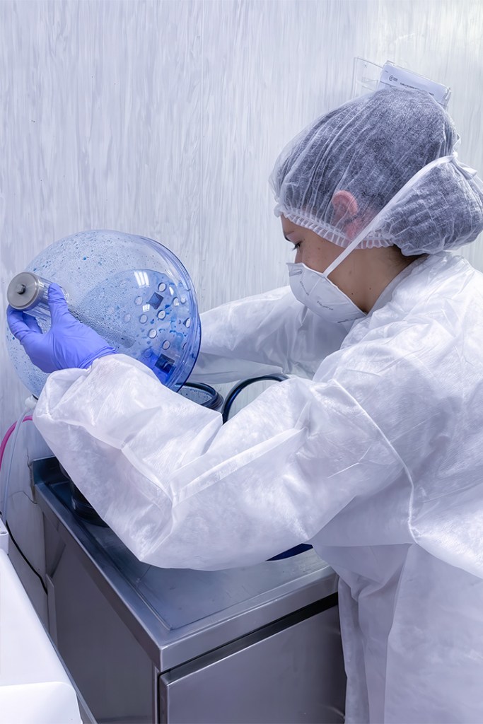 Person in protective gear handling a blue glass container in a lab setting.