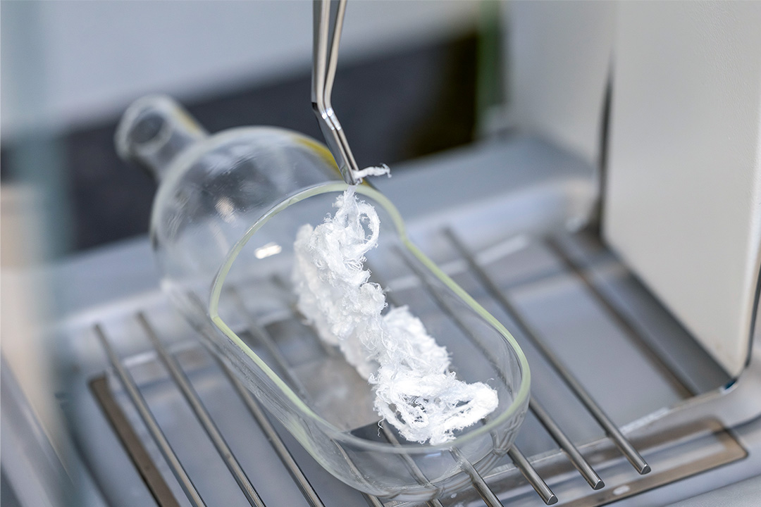 Tweezers holding white material inside a glass container on a metal rack.