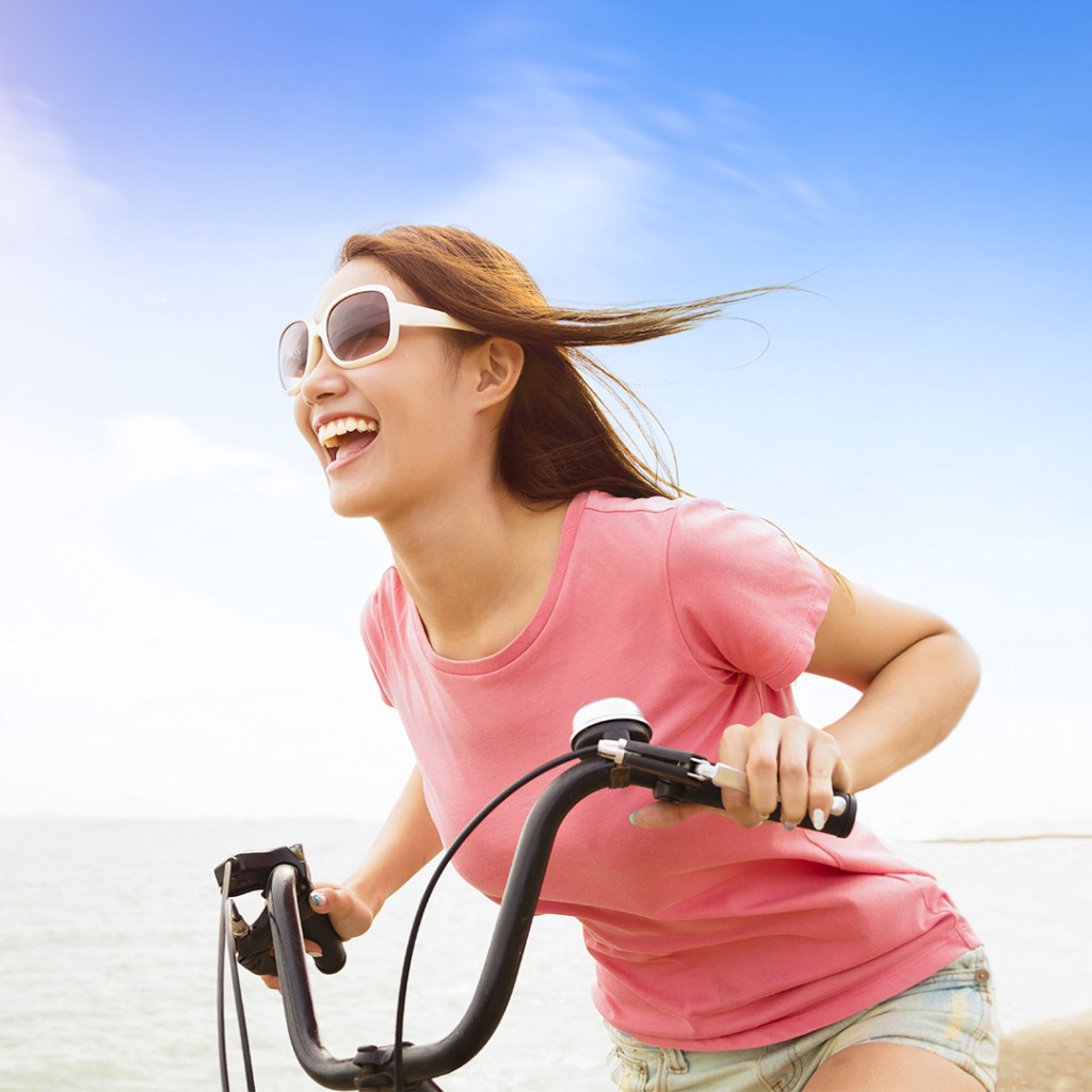 Woman in pink shirt riding a bicycle by the beach, smiling and enjoying the breeze.