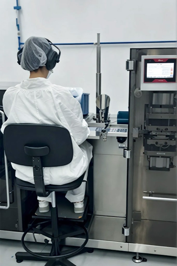Person in white lab coat and headphones operating machinery in a lab setting.